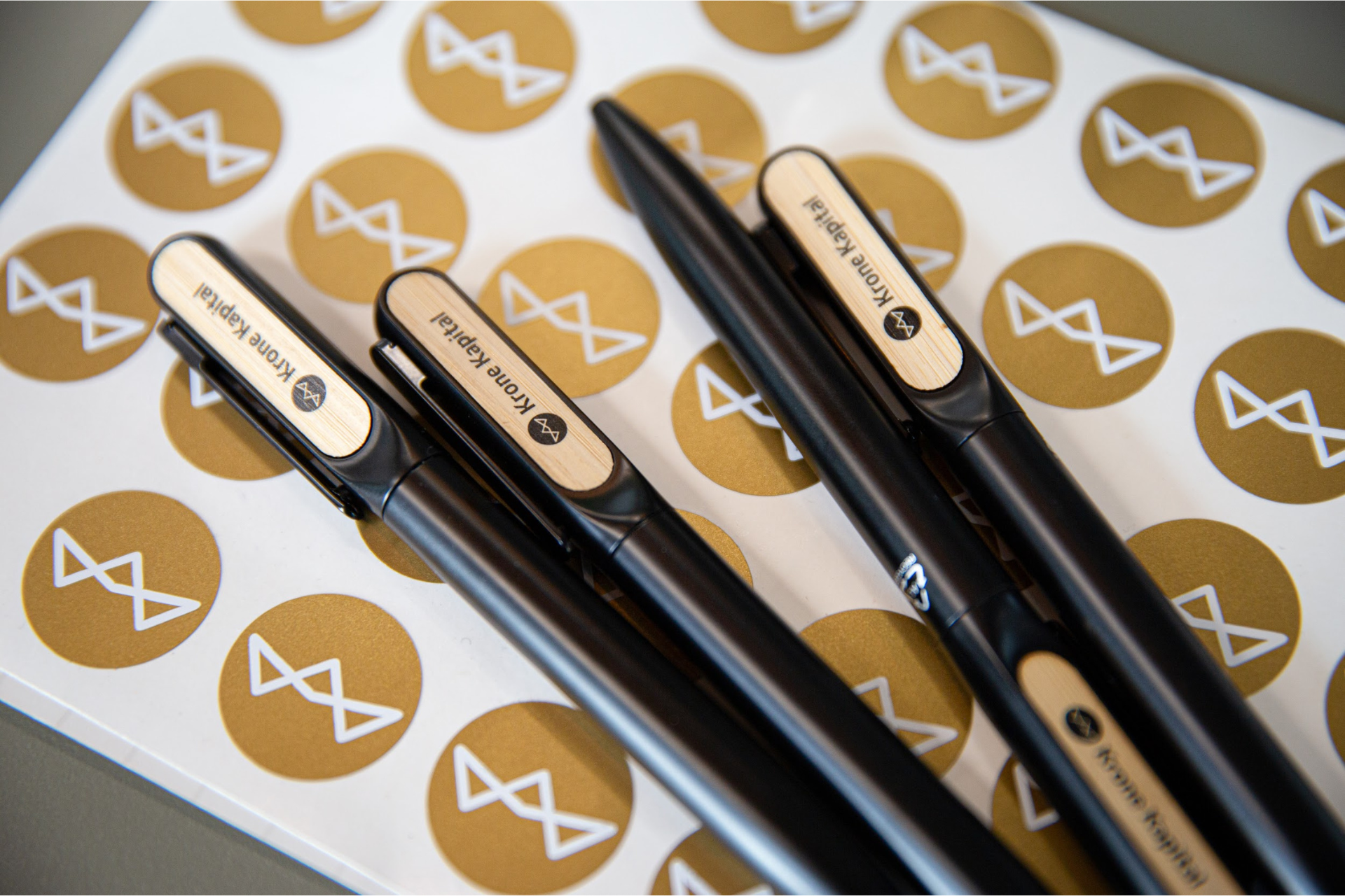 En håndfuld Krone Kapital kuglepenne ligger ovenpå klistermærke med Krone Kapital logoer i guld. Uden vandmærke.