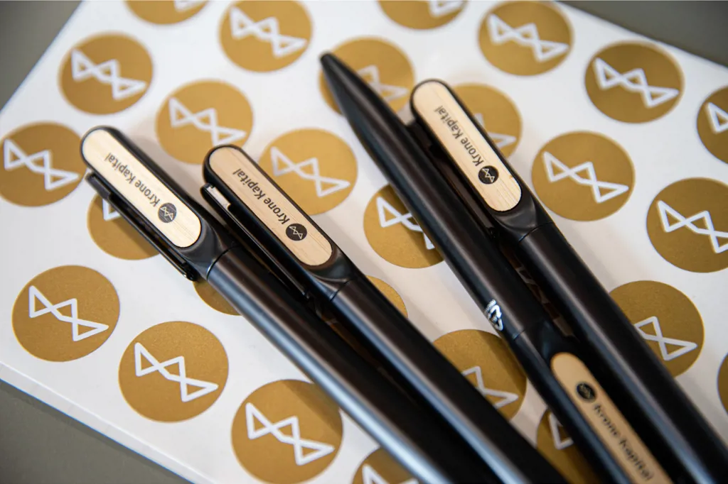 En håndfuld Krone Kapital kuglepenne ligger ovenpå klistermærke med Krone Kapital logoer i guld. Uden vandmærke.